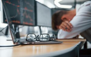 Person leaning forward on a desk with multiple computer screens in the background, appearing fatigued.