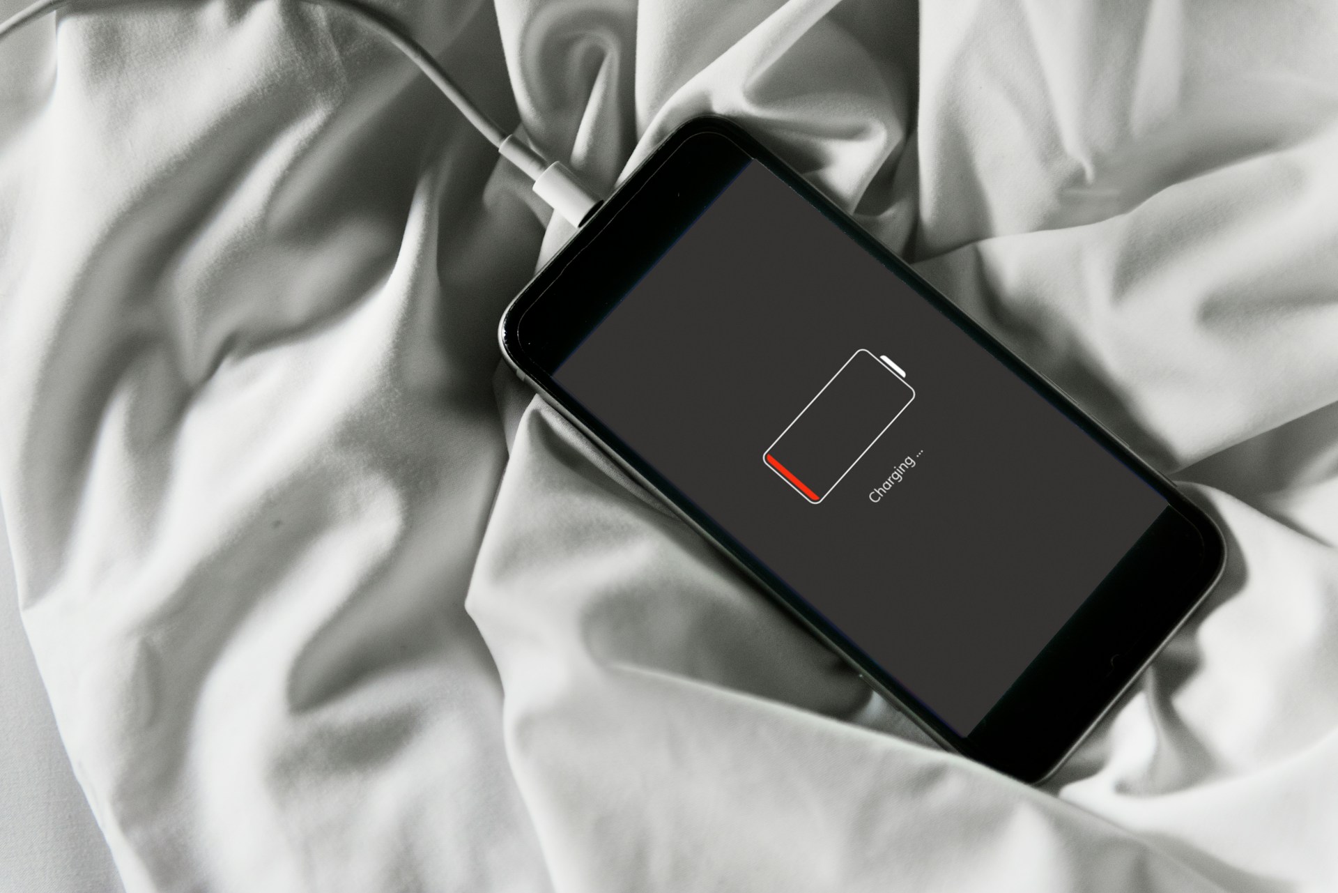 Smartphone connected to charger on white bedsheets; screen shows a battery icon and 'Charging' text.