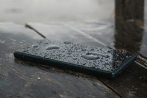Smartphone resting on a wooden surface with water droplets visible on the screen.