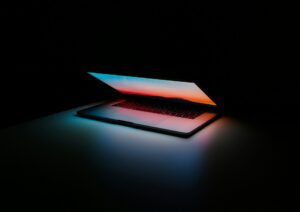 Open laptop on a desk in darkness, screen reflecting a blue-to-orange sunset gradient.