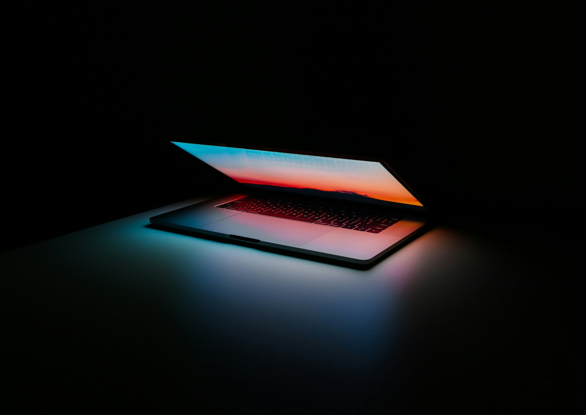 Open laptop on a desk in darkness, screen reflecting a blue-to-orange sunset gradient.