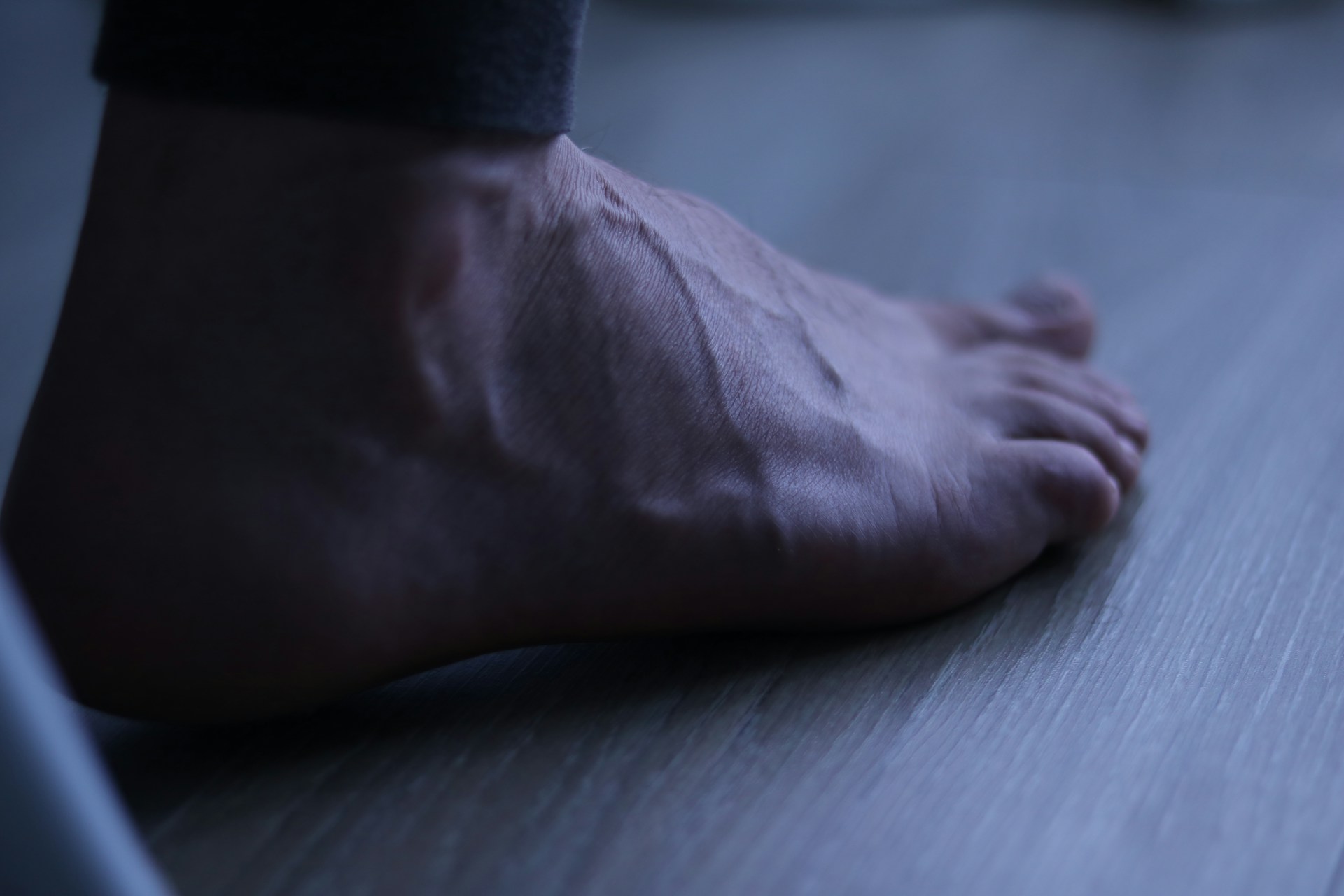 Bare foot resting on a wooden floor beside a bed at night.